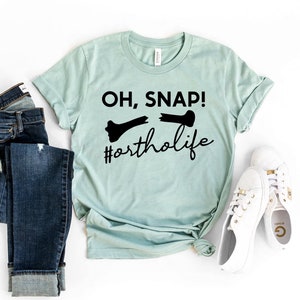 May include: A light blue t-shirt with black text that reads "OH, SNAP! #ortholife" and a broken bone graphic.