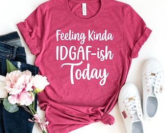 May include: A magenta t-shirt with the text "Feeling Kinda IDGAF-ish Today" in white. The shirt is styled with a pair of white sneakers, blue jeans, and a bouquet of pink and white flowers. The shirt is made of a soft material.