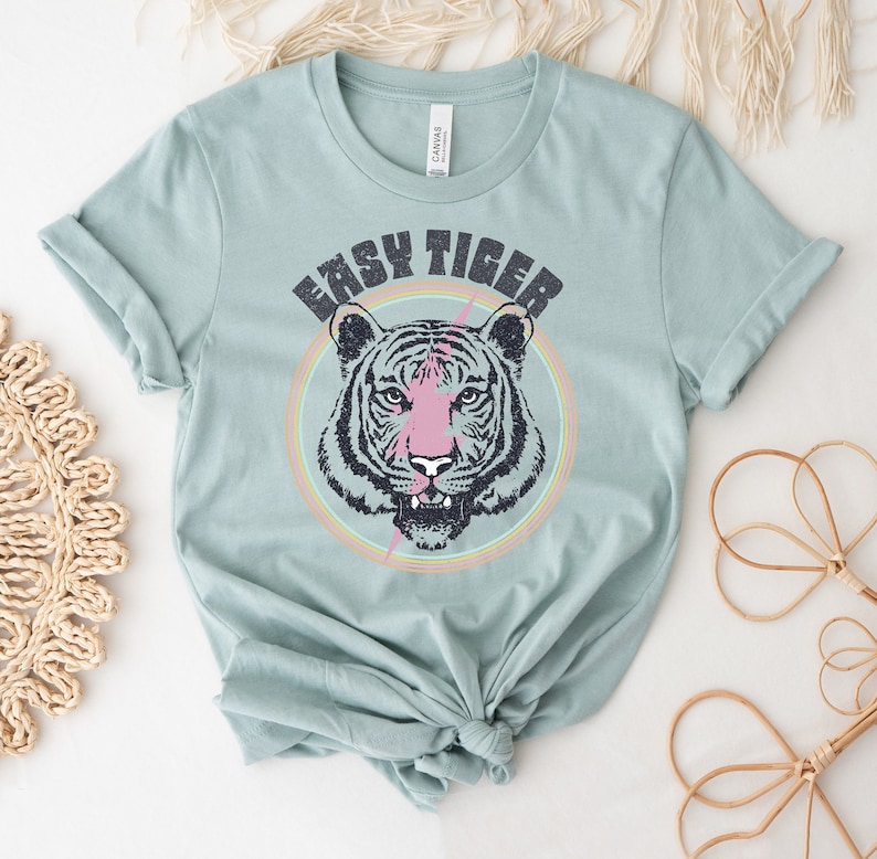 May include: A light blue t-shirt with a graphic of a tiger's face in a circle with a pink lightning bolt and the text "Easy Tiger".