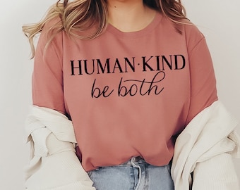 May include: A woman wearing a dusty rose long-sleeved t-shirt with the text "HUMAN KIND be both" printed in black. She is wearing blue jeans and a white cardigan.
