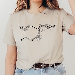 Puede incluir: Una camiseta beige con un diseño floral negro de una molécula de dopamina con la palabra "Dopamina" escrita en cursiva.