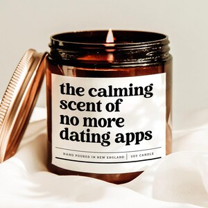 May include: A lit soy candle in a brown glass jar with a copper lid. The label reads "the calming scent of no more dating apps." The candle is hand-poured in New England.