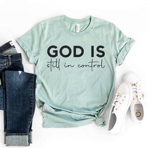 Puede incluir: Una camiseta verde claro con el texto negro "GOD IS still in control". La camiseta se combina con vaqueros azules y zapatillas blancas.