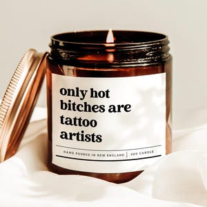 May include: A lit soy candle in a brown glass jar with a gold lid. The label reads "only hot bitches are tattoo artists" in black text. The candle is hand-poured in New England.