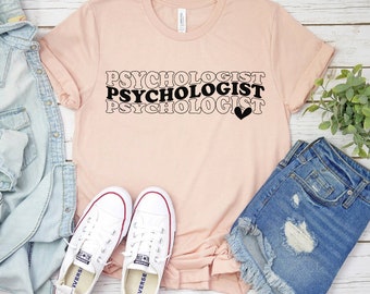 May include: A light pink t-shirt with the word "PSYCHOLOGIST" in a stacked design, with a small black heart. The shirt is paired with white sneakers, denim shorts, and a denim jacket.
