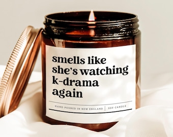 Może przedstawiać: Zapalone świeca sojowa w brązowym szklanym słoiku ze złotą pokrywką. Etykieta głosi "smells like she's watching k-drama again". Świeca jest ręcznie wylewana w Nowej Anglii.
