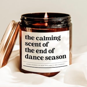 May include: A lit soy candle in a brown glass jar with a copper lid. The label reads "the calming scent of the end of dance season." The candle is hand-poured in New England.