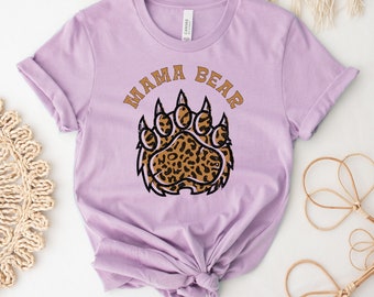 Puede incluir: Una camiseta lavanda con el texto "Mama Bear" en letras marrones y una huella de pata de oso marrón con un diseño de estampado de leopardo.