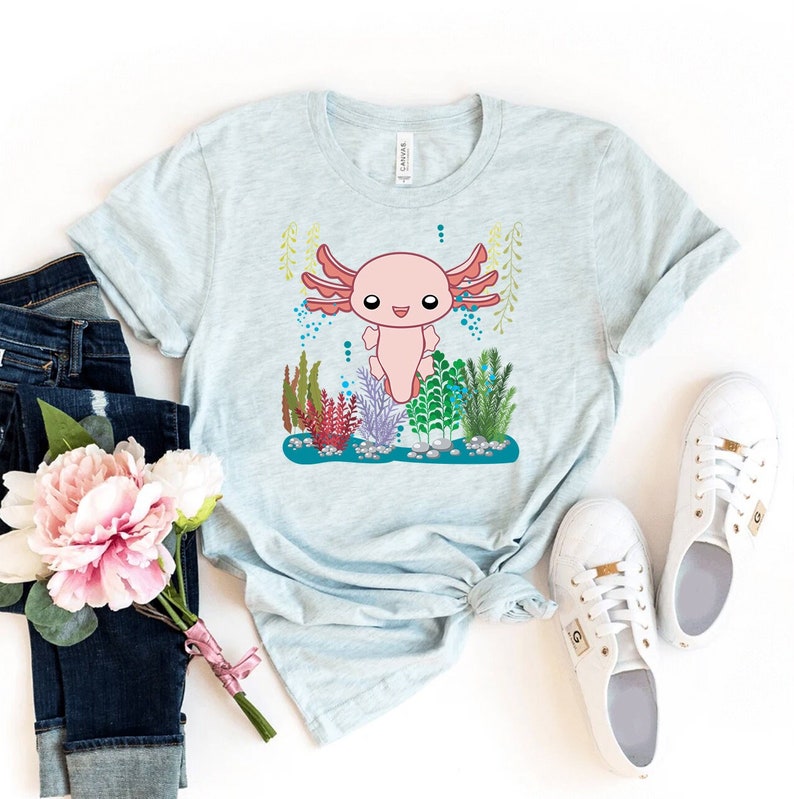 K&ouml;nnte beinhalten: Ein hellblaues T-Shirt mit einem Cartoon-Axolotl in einem rosa-roten Farbschema. Der Axolotl ist von gr&uuml;nen, blauen und roten Wasserpflanzen umgeben. Das T-Shirt liegt flach auf einer wei&szlig;en Oberfl&auml;che.