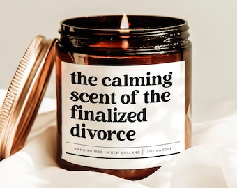 May include: A lit soy candle in a brown glass jar with a gold lid. The label reads "the calming scent of the finalized divorce." The candle is hand-poured in New England.