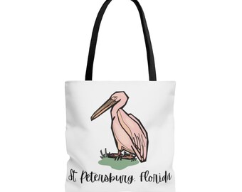 May include: White tote bag with a pink pelican illustration and black handles. The pelican stands on green grass, with the text "St. Petersburg, Florida" in black script below. The bag is suitable for carrying items.