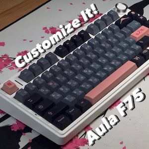 May include: A white Aula F75 mechanical keyboard with black and gray keycaps. The keyboard has a pink and white space bar and a pink escape key. The keyboard is on a white mousepad with a pink cherry blossom design. The text "Customize It!" is written in white on a black background above the keyboard.