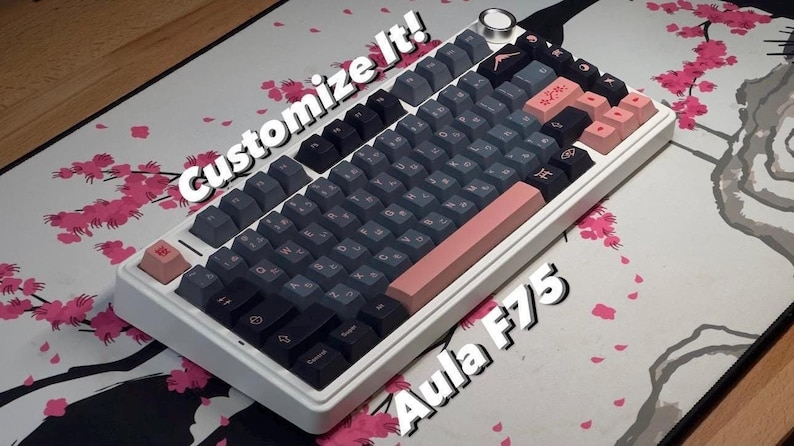 May include: A white Aula F75 mechanical keyboard with black and gray keycaps. The keyboard has a pink and white space bar and a pink escape key. The keyboard is on a white mousepad with a pink cherry blossom design. The text "Customize It!" is written in white on a black background above the keyboard.