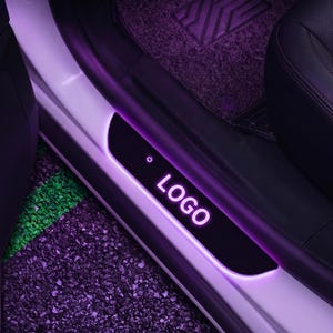 May include: A car door sill plate with the word "LOGO" illuminated in purple. The plate is black with a purple glow around the edges. A power symbol is visible on the left side of the plate.