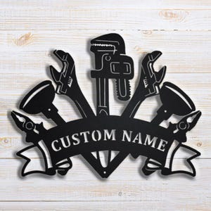 Peut inclure: Décoration murale en métal noir représentant des outils de plomberie, dont une clé à molette, des clés, des pinces et des ventouses. Une bannière avec l'inscription "CUSTOM NAME" en lettres blanches. Sur fond de bois clair.