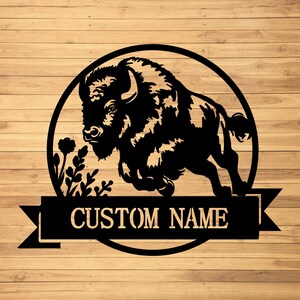 May include: Black metal bison wall art with a circular frame, floral accents, and a banner that reads "CUSTOM NAME." The bison is depicted in a running pose. The background is a wood grain pattern.