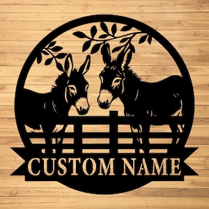 May include: Black metal wall art depicting two donkeys behind a fence, framed by a circular design with leaves. The words "CUSTOM NAME" are displayed on a banner below the fence. The background has a wood grain pattern.