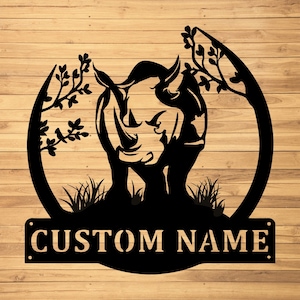 May include: Black metal rhino silhouette with a custom name plate. The rhino is framed by stylized trees and grass. The text "CUSTOM NAME" is displayed on a rectangular plate at the bottom. The background is a wood grain pattern.
