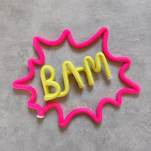 Pode incluir: Um círculo de fio rosa brilhante com a palavra "BAM" em fio amarelo dentro. O círculo de fio tem a forma de uma explosão de banda desenhada.