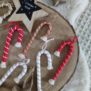Può includere: Cinque decorazioni di bastoncino di zucchero fatte a mano con strisce rosse e bianche e glitter dorato. Le decorazioni sono appese a una fetta di legno con un cartello a forma di stella che dice "Joyeux Noel".