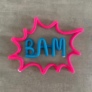 Pode incluir: Um círculo de fio rosa com a palavra "BAM" em fio azul dentro.