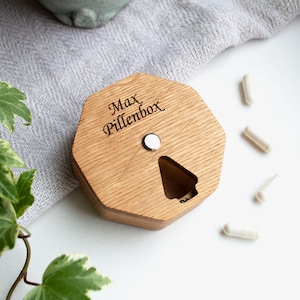 May include: Wooden pill box with the engraved text "Max Pillenbox". The hexagonal box has a magnetic closure and a compartment opening. Several white capsules are scattered nearby. Green and white ivy leaves are in the corner.