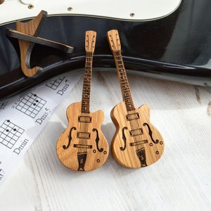 Könnte beinhalten: Zwei Miniatur-Holzgitarren mit einem schwarz-weißen Gitarrenakkord-Diagramm im Hintergrund. Die Gitarren sind mit Bundlinien und einem Tonabnehmer detailliert.