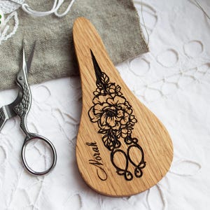 Op de afbeelding: Een houten scharenstandaard met een bloemenontwerp en de naam "Norah" in zwart geëtst. Een zilveren borduurschaar en een bloemenvingerhoed liggen op een wit oppervlak met een grijze stof.