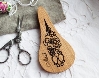 May include: A wooden scissor holder with a floral design and the name "Norah" etched in black. A pair of silver embroidery scissors and a floral thimble rest on a white surface with a gray fabric.