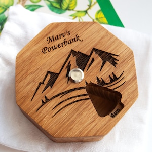 May include: Wooden power bank with a mountain design. The octagonal, light brown power bank has the text "Marv's Powerbank" and a mountain range etched into the wood. A silver button and a triangular cutout are visible.