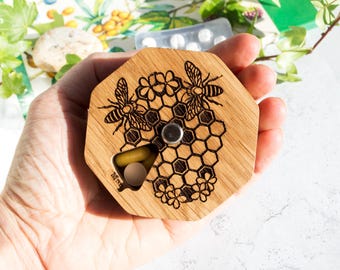 Peut inclure: Boîte à pilules en bois de forme hexagonale avec un motif en nid d'abeilles. Le couvercle est gravé de deux abeilles et de fleurs. La boîte est ouverte et montre deux pilules à l'intérieur.