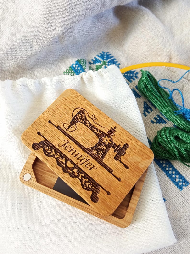 A handcrafted wooden needle storage box with an engraved vintage sewing machine design and the name "Jennifer" inscribed. The box is placed on a linen cloth with embroidery, accompanied by green embroidery floss and a yellow embroidery hoop