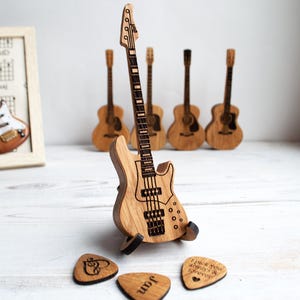 Może przedstawiać: Drewniana figurka gitary basowej stoi przed kilkoma figurkami gitary akustycznej. Gitara basowa jest jasnobrązowa z wytrawionymi detalami. Trzy drewniane kostki do gitary z wytrawionymi wzorami i tekstem znajdują się przed gitarą basową.