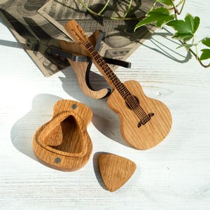 Könnte beinhalten: Holzaccessoires in Gitarrenform, darunter eine Miniaturgitarre, ein Plektrum und ein Etui. Die Gitarre steht auf einem Holzständer. Die Artikel sind aus hellem Holz und liegen auf einer weißen Oberfläche.