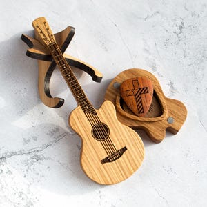 Puede incluir: Una guitarra de madera con un soporte para púa. La guitarra está hecha de madera clara y tiene un soporte para púa de madera oscura. El soporte para púa tiene un cierre magnético y una púa con un diseño de cruz en su interior.