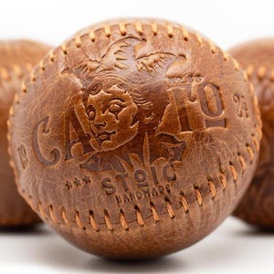 Może przedstawiać: Trzy brązowe skórzane piłki baseballowe ze szwami. Piłka środkowa ma wytłoczony tekst "CASTLE" i "STOIC HANDMADE".
