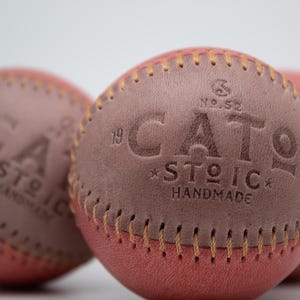 Op de afbeelding: Close-up van drie handgemaakte honkballen. De honkballen zijn rood en bruin met zichtbare stiksels. De tekst "CATO STOIC HANDMADE" is in het bruine leer geëmbosseerd. De honkballen zijn scherp gesteld tegen een witte achtergrond.