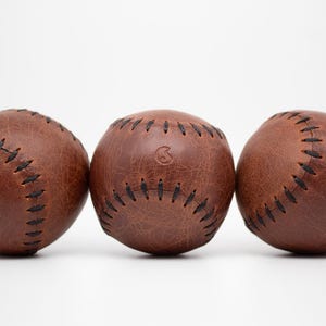 Op de afbeelding: Drie bruine leren honkballen met zwarte stiksels zijn gerangschikt tegen een witte achtergrond. De honkballen hebben een versleten, vintage uitstraling, met zichtbare nerf en textuur. Eén bal heeft een logo.