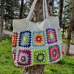 May include: A white crocheted tote bag with colorful granny square patches. The bag has a long strap and is hanging on a tree trunk.