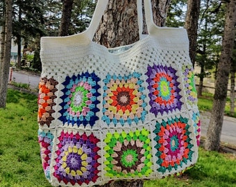 May include: A white crocheted tote bag with colorful granny square patches. The bag has a long strap and is hanging on a tree trunk.
