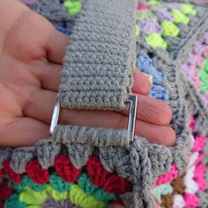 Op de afbeelding: Een grijze gehaakte tas met een kleurrijk granny square patroon. De tas heeft een grijze gehaakte band met een zilveren metalen gesp.
