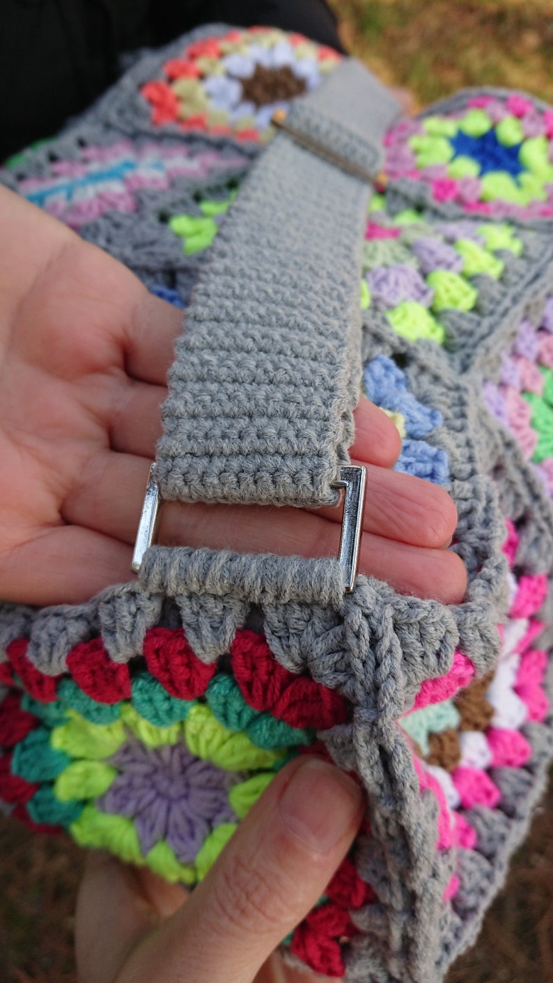 Op de afbeelding: Een grijze gehaakte tas met een kleurrijk granny square patroon. De tas heeft een grijze gehaakte band met een zilveren metalen gesp.