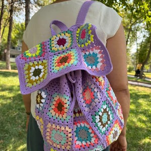 May include: A purple crochet backpack with a drawstring closure. The backpack is made of granny squares with colorful flowers in the center of each square.