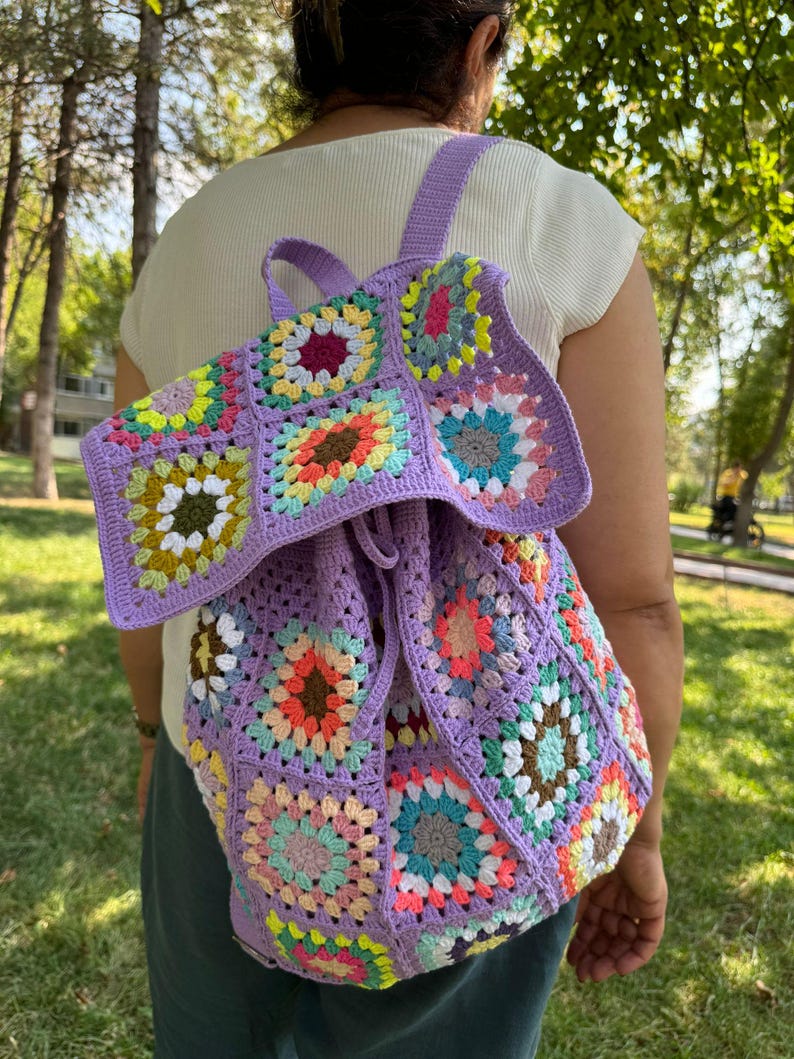 May include: A purple crochet backpack with a drawstring closure. The backpack is made of granny squares with colorful flowers in the center of each square.