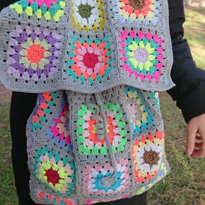 Op de afbeelding: Een grijze gehaakte rugzak met kleurrijke granny squares. De rugzak heeft een trekkoord sluiting en een bovenste handvat.