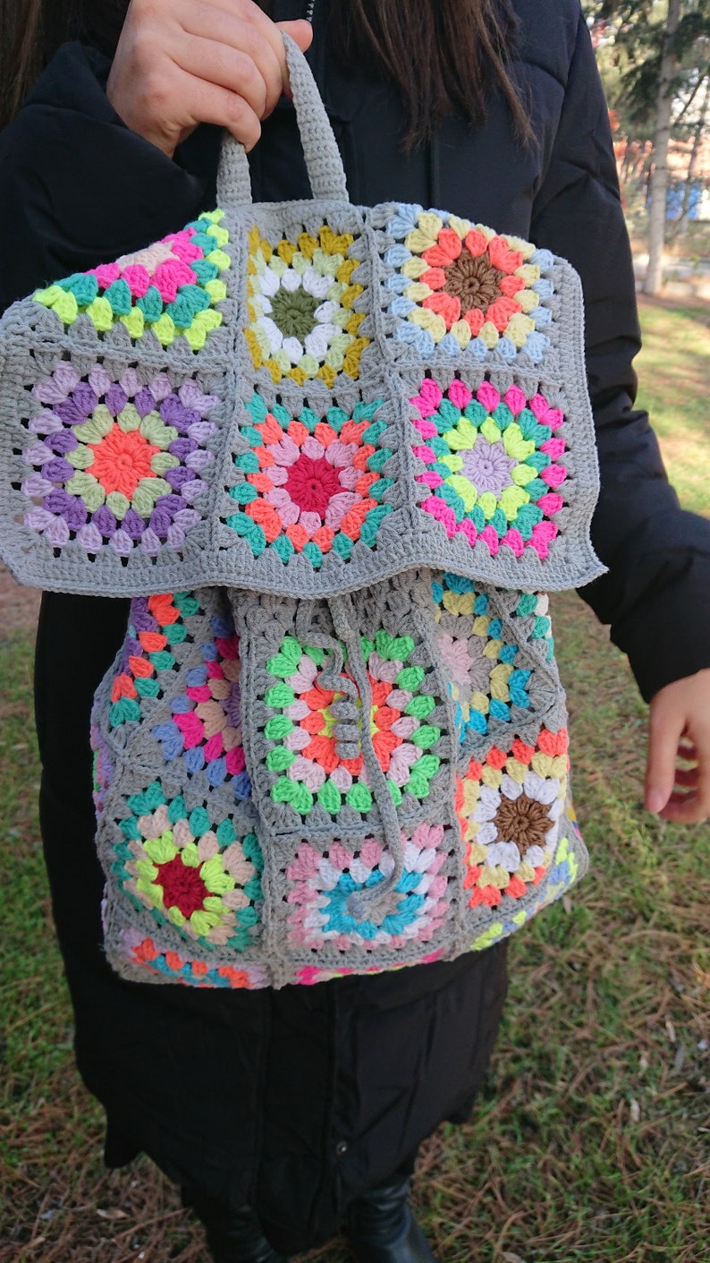 Op de afbeelding: Een grijze gehaakte rugzak met kleurrijke granny squares. De rugzak heeft een trekkoord sluiting en een bovenste handvat.