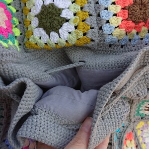 Op de afbeelding: Een grijze gehaakte tas met een kleurrijk granny square patroon. De tas is open om een grijze voering te laten zien.