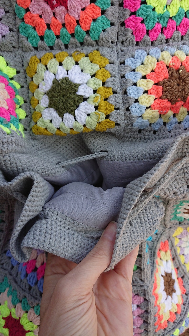 Op de afbeelding: Een grijze gehaakte tas met een kleurrijk granny square patroon. De tas is open om een grijze voering te laten zien.