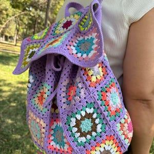 May include: A purple crochet backpack with a drawstring closure. The backpack is made of granny squares in various colors, including pink, green, yellow, and brown.