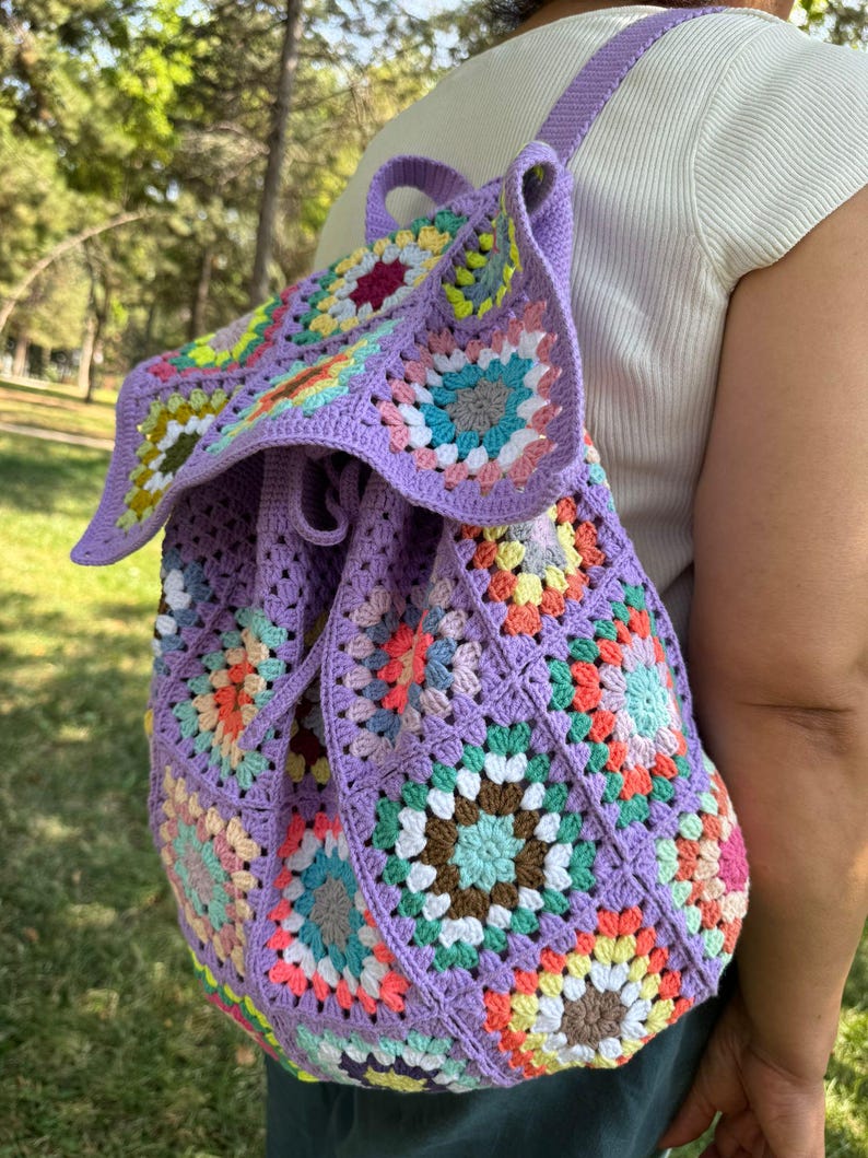 May include: A purple crochet backpack with a drawstring closure. The backpack is made of granny squares in various colors, including pink, green, yellow, and brown.